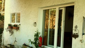 Una casa con porche adoquinado, ventana con vistas a árboles y puerta con felpudo rojo.