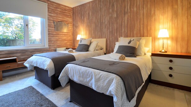 A bedroom with two beds, wooden paneling on the walls, bedside lamps, and a window with a view of trees. - Cedar Cottage at Rashfield nr Dunoon-sleeps up to 6-Pet Friendly(1 max) (Dunoon)