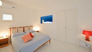 Ein Schlafzimmer mit Bett, Nachttischen, einer Wand-Klimaanlage, einem Fenster mit Jalousien, einer Tür und einem Wandbild.