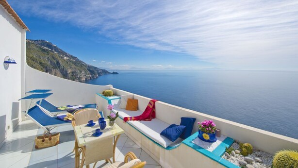 Ein Balkon mit weißer Wand, blauen Kissen, einem Tisch mit Blumenvasen und Blick auf das Meer und die Klippen.