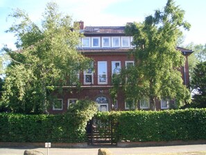 Un edificio de ladrillo de dos pisos con un diseño tradicional, que cuenta con una entrada central con portón, rodeado de exuberante vegetación.