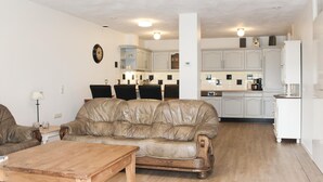 Una sala de estar con un sofá de cuero, una mesa de centro de madera, y un comedor con sillas, con una cocina al fondo.