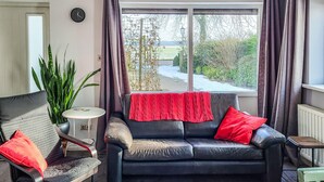 A cozy living room with a black leather sofa, a small round table, and a chair. There is a large window with curtains, a potted plant, and a view of a pathway and greenery outside. - 1 bedroom cozy home in Sprundel (Sprundel)