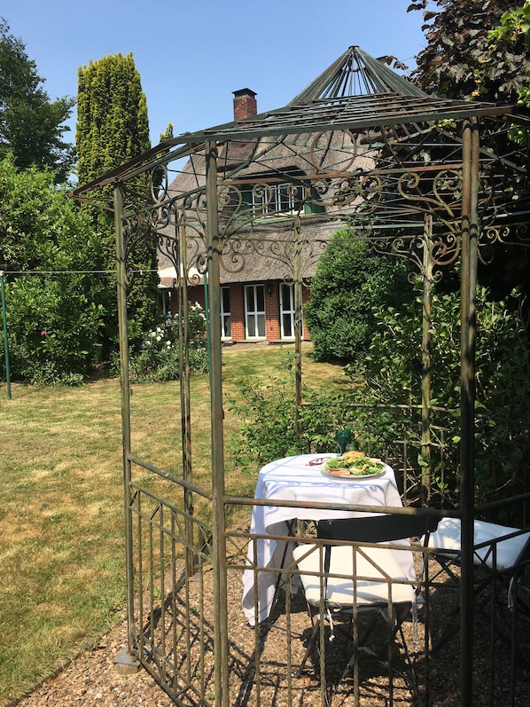 Relax under the gazebo, surrounded by lush gardens and a charming brick building in the background.