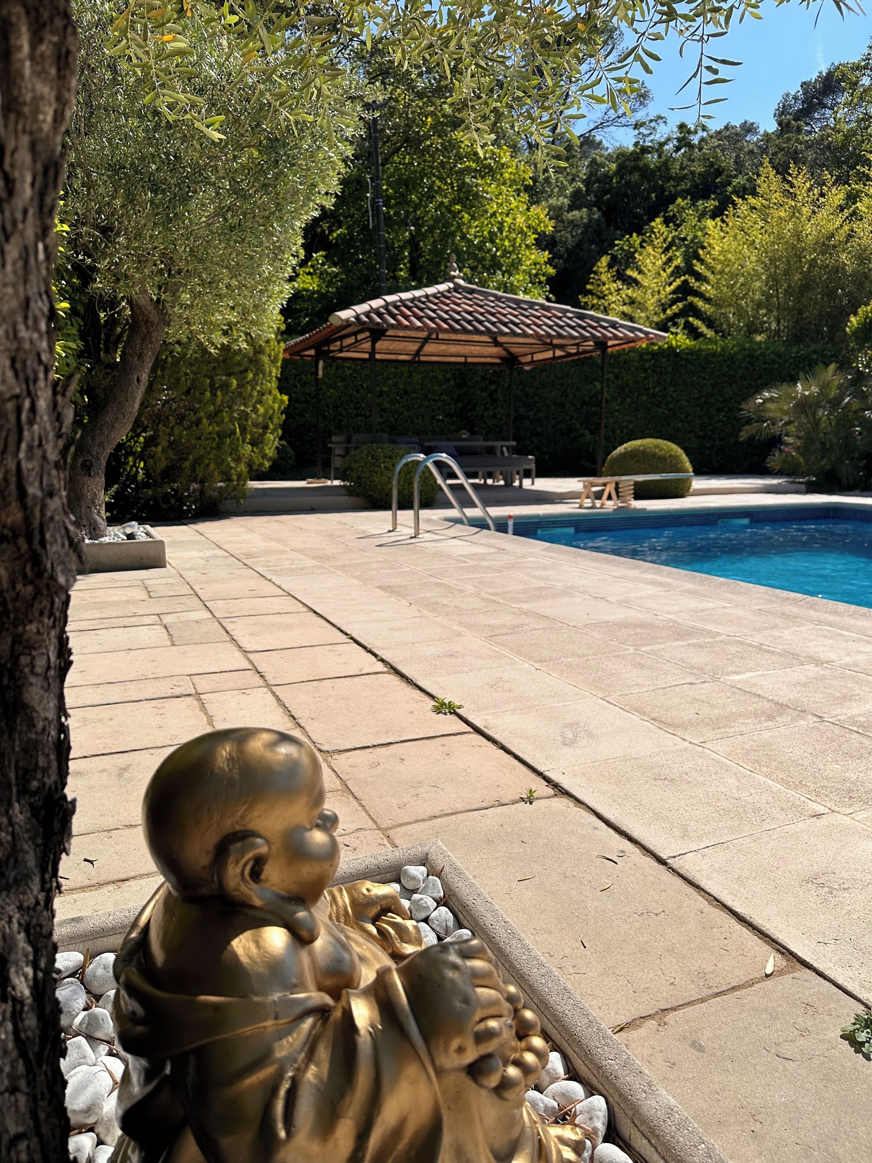 Una estatua de una persona en un jardín, con una piscina y una pérgola al fondo.