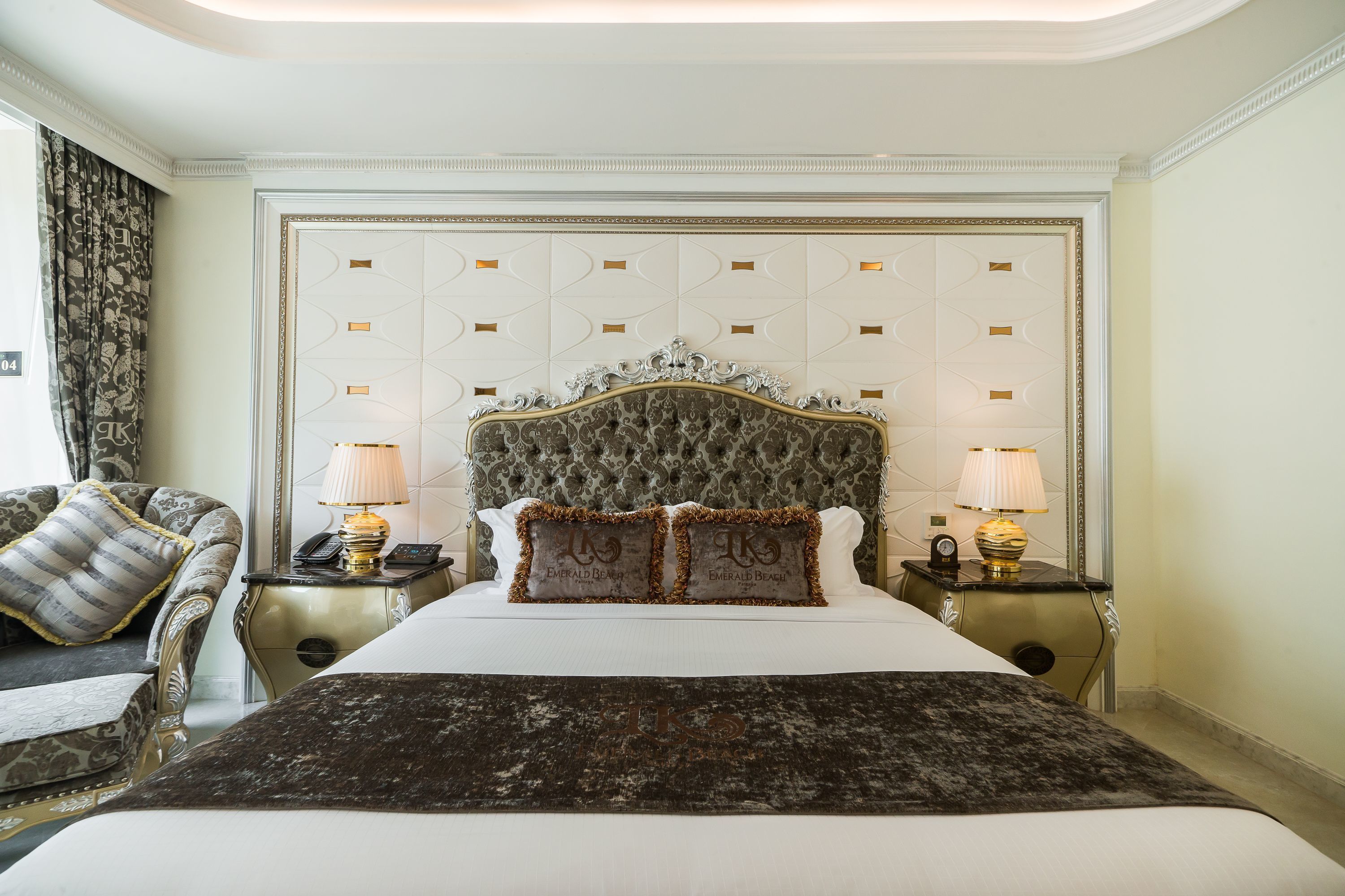 Un dormitorio lujoso con una cama grande, un cabecero decorativo, dos mesitas de noche con lámparas y un sillón clásico.