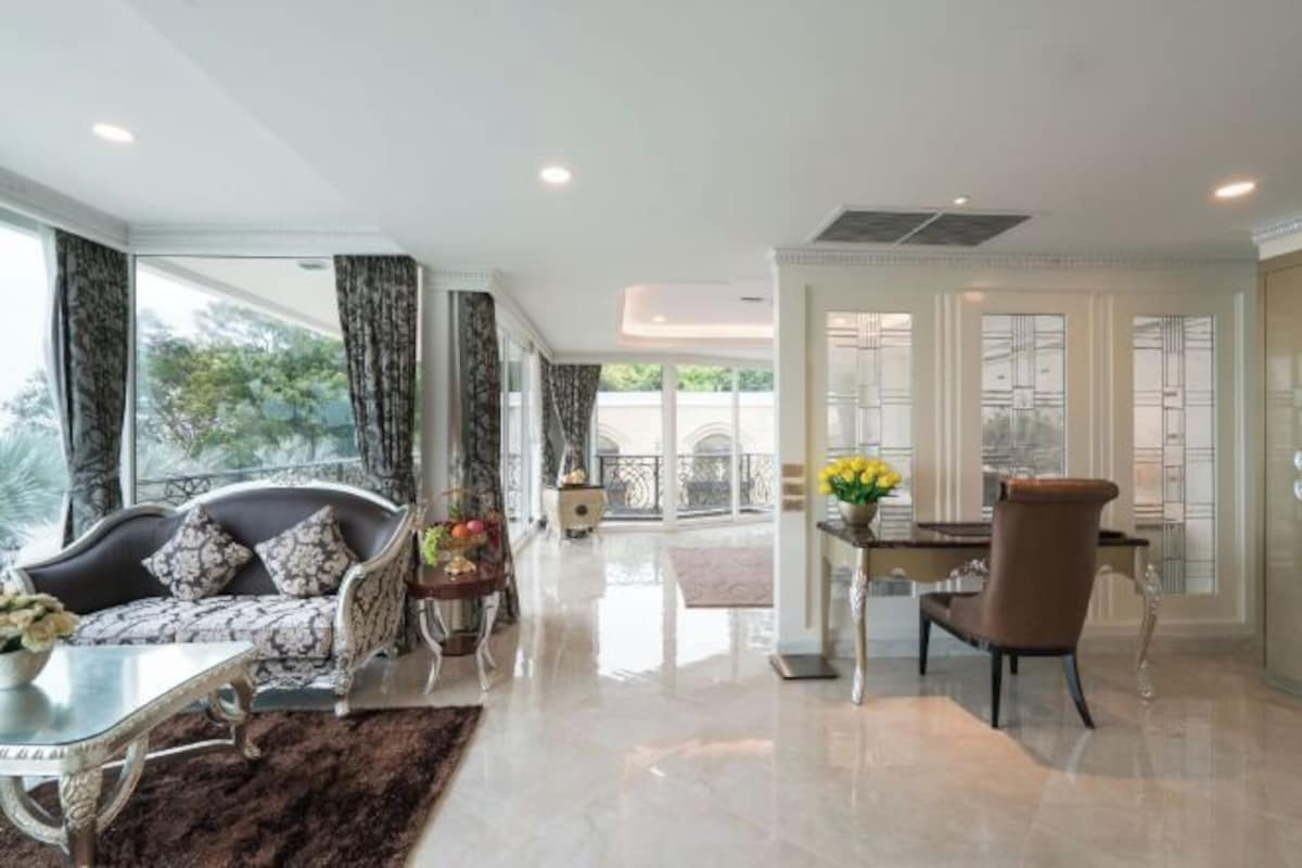 A spacious living room with a sofa, a dining table, and a chair. There is a balcony with a railing and a view of trees.