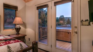 A bedroom with a wooden ceiling, a bed with a red and white blanket, a lamp, and a view of a deck and trees through a sliding door.