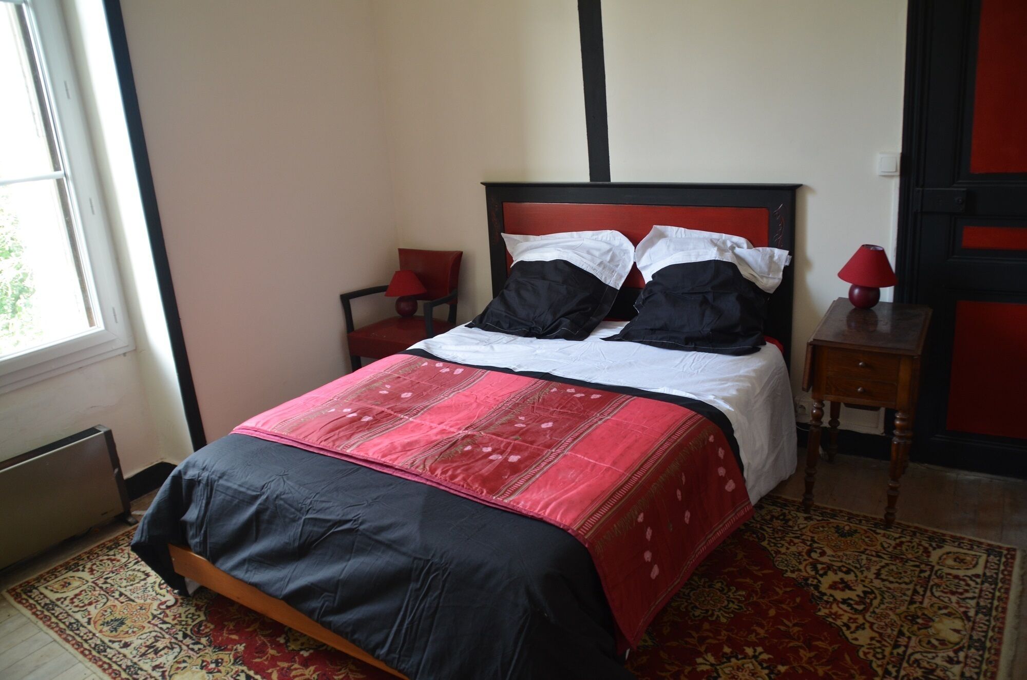 Un lit bien fait, recouvert d’une couvre-lit rouge et noire, avec une tête de lit en bois et une lampe rouge posée sur une table de chevet.
