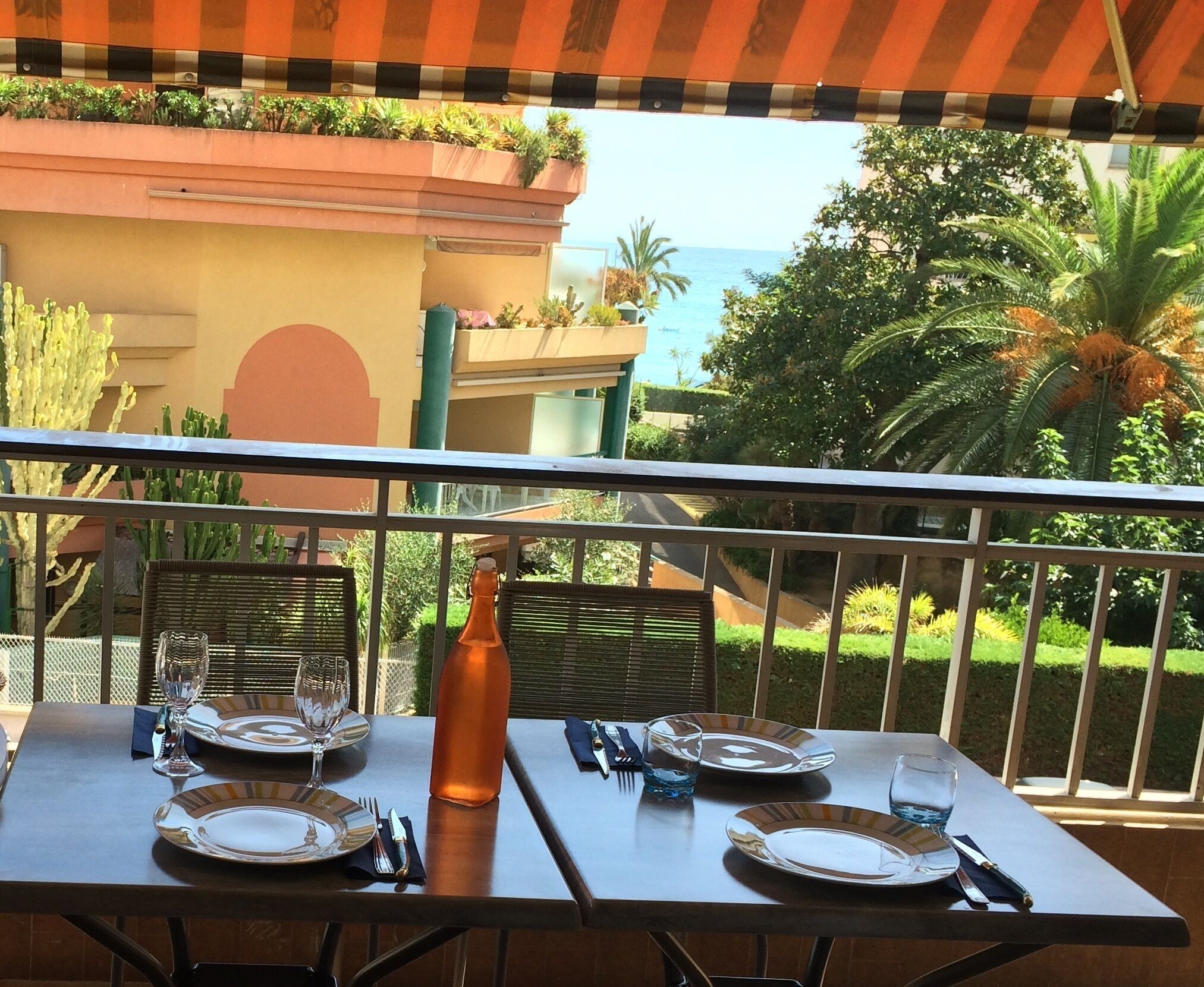 A dining table set for two with plates, cutlery, and a bottle, overlooking a garden and a building with a view of the sea.