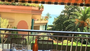 A dining table set for two with plates, cutlery, and a bottle, overlooking a garden and a building with a view of the sea. - APARTMENT 6 pers 150m SEASIDE SEA LARGE TERRACE SEA VIEW PARKING (Roquebrune-Cap-Martin)