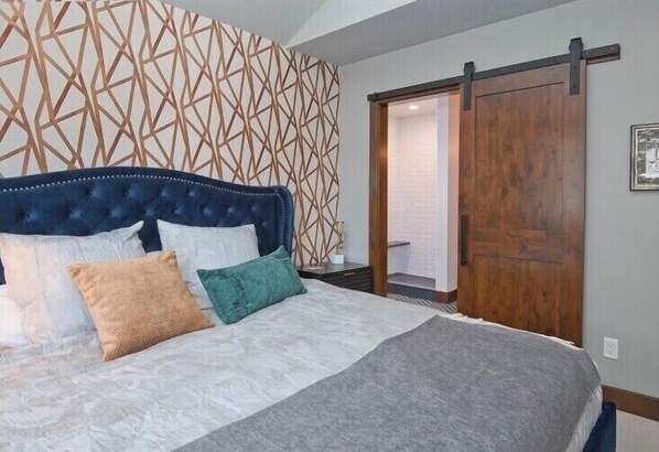 Ein Schlafzimmer mit einer gesteppten Kopfteil, einem großen Bett mit vielen Kissen, einer Wand mit geometrischem Muster und einer Holztür, die sich gleitet.