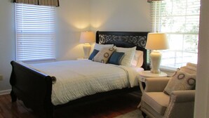 Une chambre à coucher comprenant un lit, des lampes de chevet, une chaise, un ventilateur de plafond et des fenêtres équipées de stores.