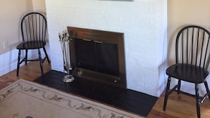 Un salon avec une cheminée en briques blanches, deux fauteuils noirs et un tableau coloré représentant une vache accroché au mur.