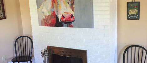 Un salón con chimenea de ladrillo blanco, dos sillones negros y un cuadro llamativo de una vaca en la pared.