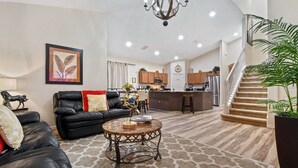 Ein geräumiges Wohnzimmer mit einer schwarzen Ledersofa, einem runden Couchtisch und einer Deckenleuchte. Im Hintergrund ist eine Küche mit Holzausbau und einem Edelstahlkühlschrank erkennbar.