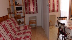 Un salon chaleureux avec un canapé, un téléviseur posé sur une étagère en bois, une table à manger avec des chaises et des rideaux à motifs de cœurs.