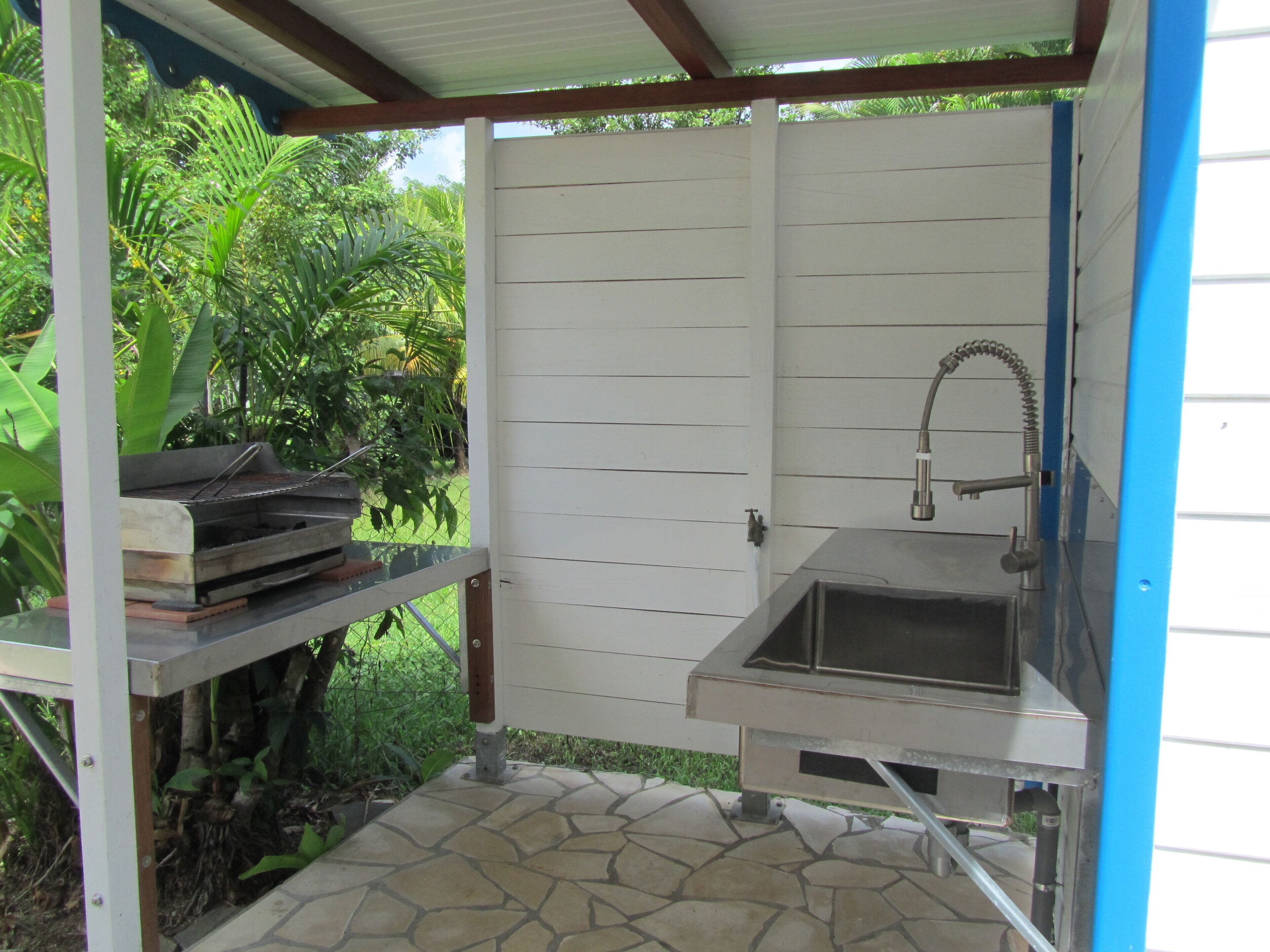 A patio area with a grill and sink, surrounded by a white fence and lush greenery.