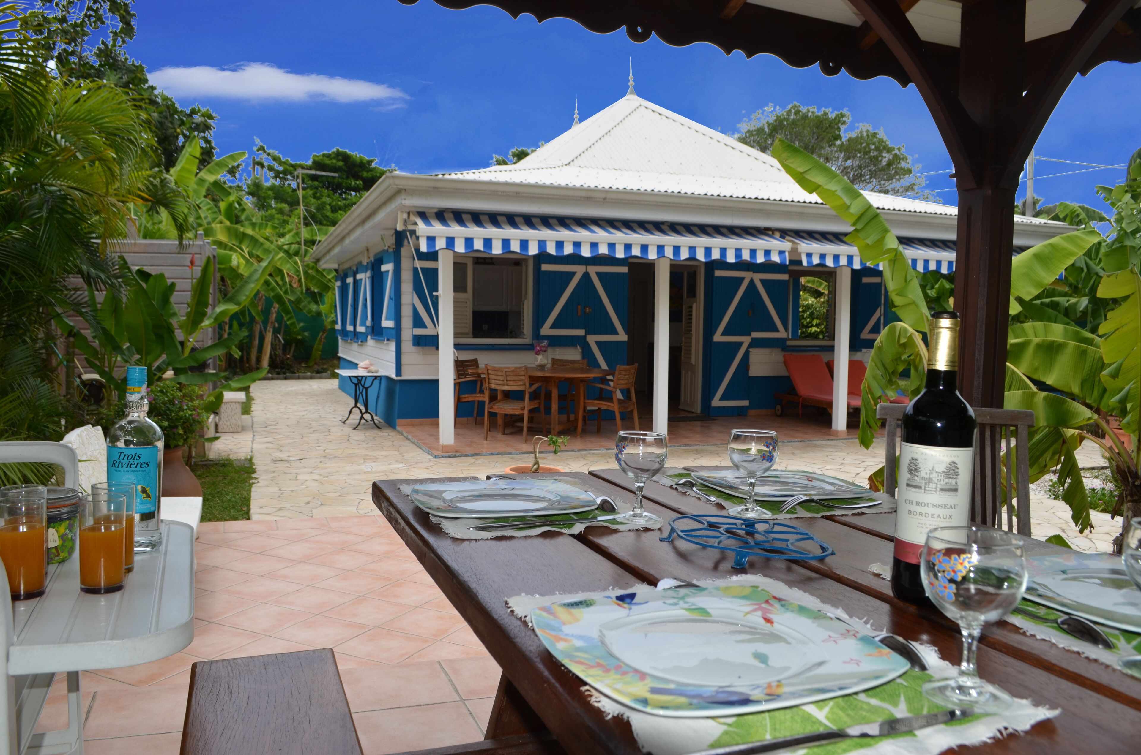 An outdoor dining area with a table set for a meal, a bottle of wine, and a tropical house in the background.