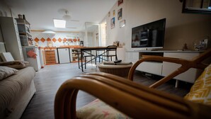 A living room with a sofa, a wicker chair, a television, and a kitchen in the background. - Cottage between Bordeaux and St Emilion with Magnificent Park, surrounded by Vines (Génissac)