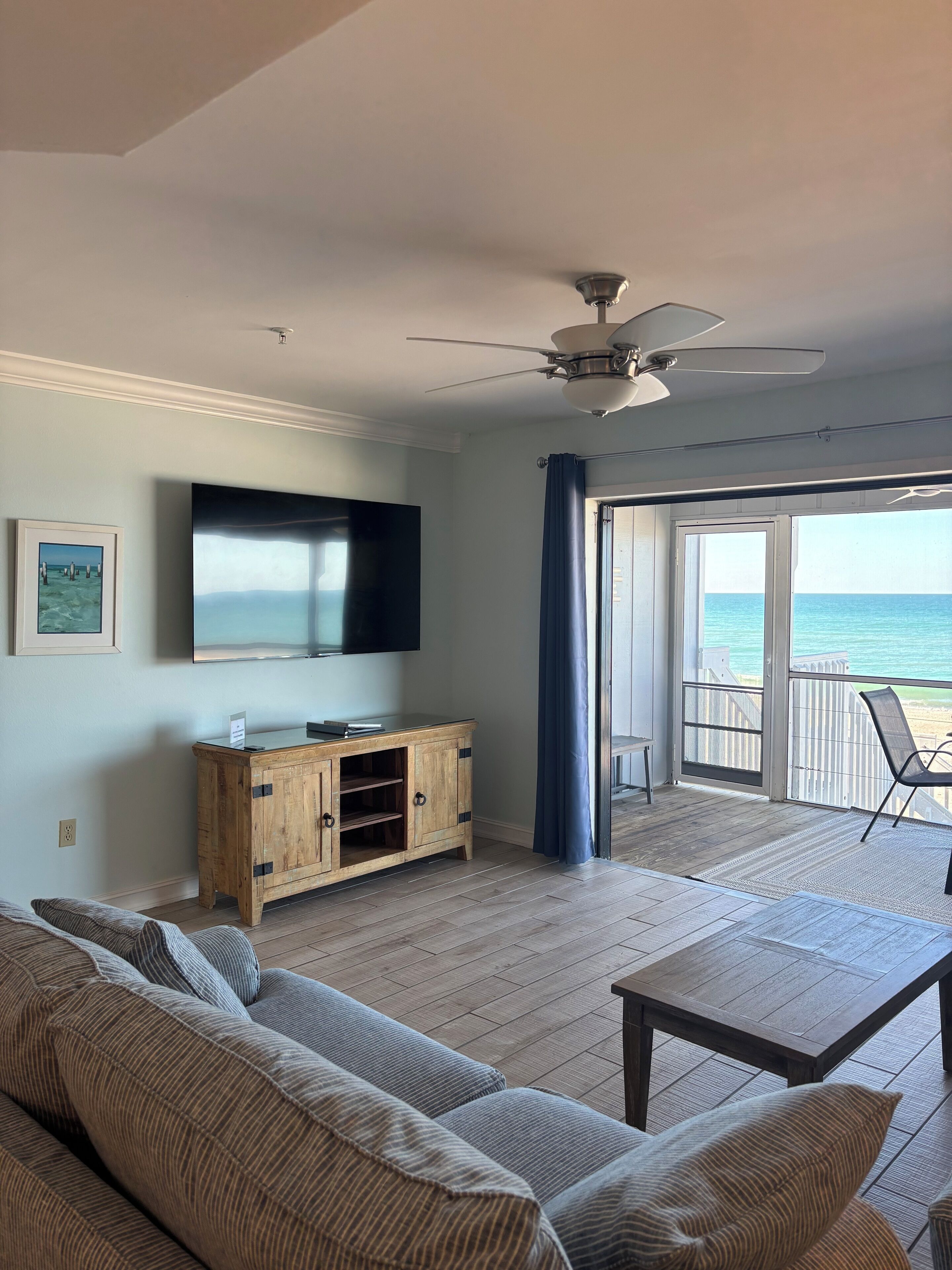 Sala de estar con un televisor grande, mueble de madera y vista al océano a través de un ventanal corredero.