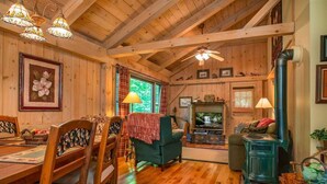 A wooden interior with a dining table, chairs, a green armchair, a fireplace, and a wood stove. - Grey Haven Cabin in Connestee Falls (Brevard)