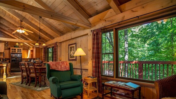 A cozy cabin interior with wooden floors, a dining area, a green armchair, a table with books, and a large window overlooking a forest. - Grey Haven Cabin in Connestee Falls (Brevard)