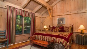 A wooden cabin bedroom with a bed, two bedside lamps, a bench, a window with curtains, and a framed picture on the wall. - Grey Haven Cabin in Connestee Falls (Brevard)