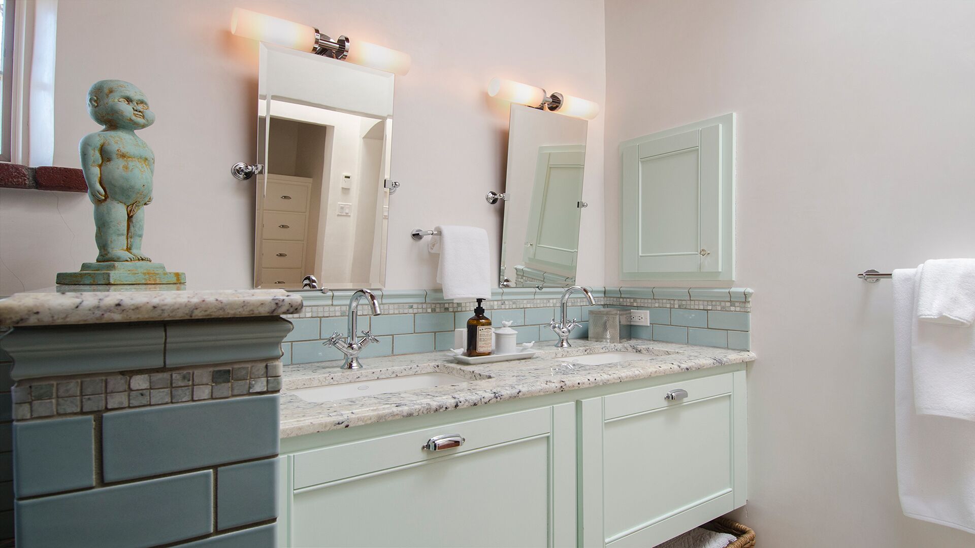 Un baño con encimera de mármol, lavamanos, espejo y una estatua.