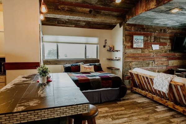 Ein gemütliches Zimmer mit Holzboden, einem Bett mit farbenfroher Steppdecke, einem kleinen Tisch mit Pflanze und einer Bank.