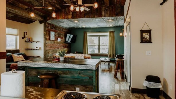 A rustic kitchen with exposed wooden beams, a brick wall, and a green island with stools. - Bridger View - Studio (Gallatin County)