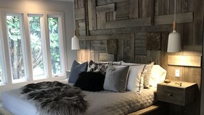 Un dormitorio moderno con cabecera de madera, una cama grande con muchos almohadones, una mesita de noche con lámpara y vistas a árboles por la ventana.
