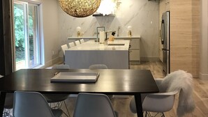 Una cocina moderna con una isla central, una zona de comedor con una mesa y sillas oscuras, y una lámpara colgante grande.