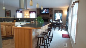 A modern kitchen with a central island, bar stools, a dining area with a brick fireplace, and a TV mounted on the wall.
