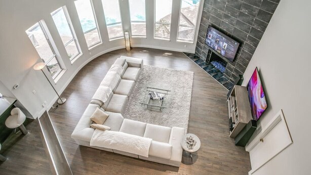 A modern living room with a large L-shaped sectional sofa, a flat-screen TV mounted on a dark wall, and a large window with a view of the outdoors. - The Best Dallas Has To Offer. Heated Pool/Spa! (Dallas)