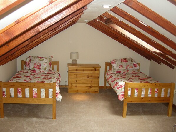 Une petite chambre mansardée avec deux lits en bois, une commode en bois et une lampe.