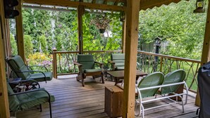 Una terraza de madera con sillas verdes y una mesa, rodeada de abundante follaje y una valla de madera.