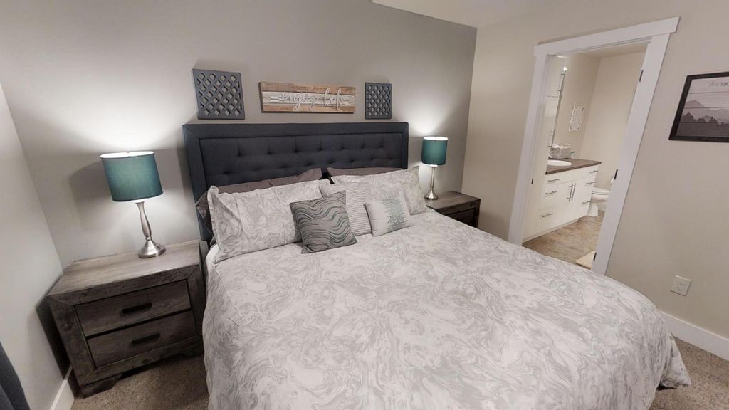 A bedroom with a large bed, two bedside tables with lamps, a dark headboard, and a door leading to a bathroom.