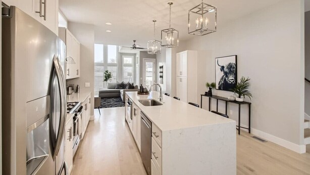Een moderne keuken met een groot kookeiland, roestvrijstalen apparatuur, hanglampen en uitzicht op de woonkamer.