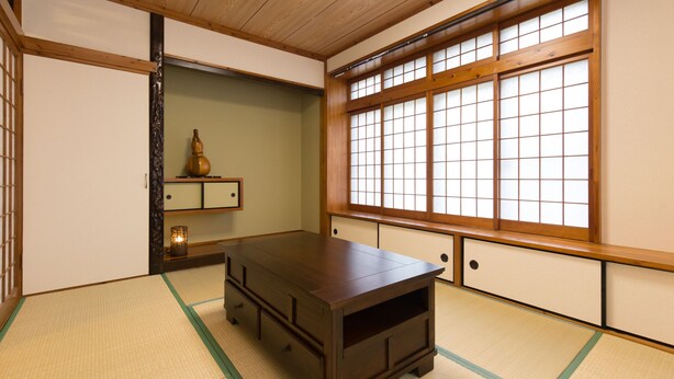 Una tradizionale stanza giapponese con pavimento in tatami, un tavolino in legno, porte scorrevoli e un'ampia finestra con infissi in legno.