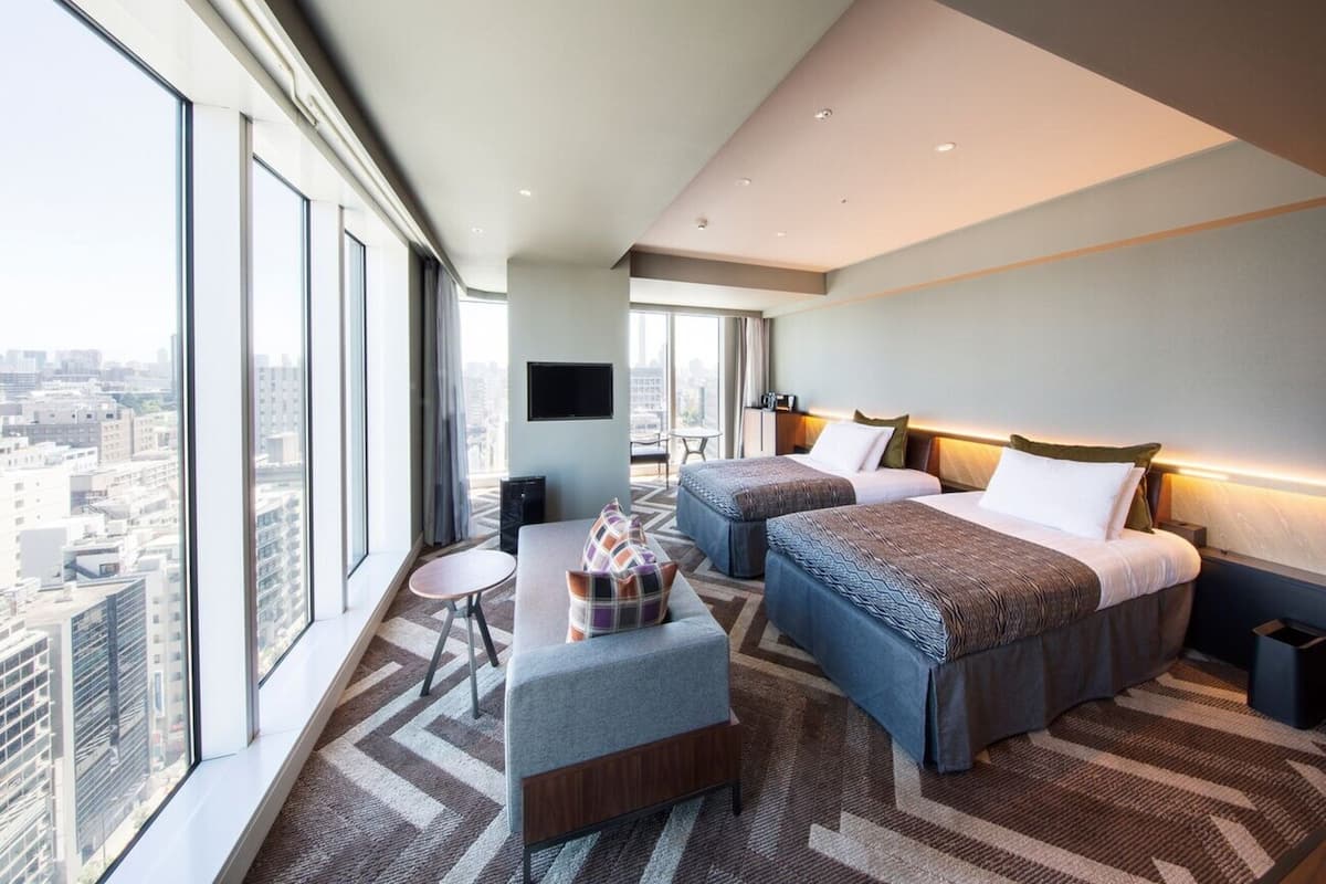 A modern hotel room with two beds, a sofa, a TV, and a city view.