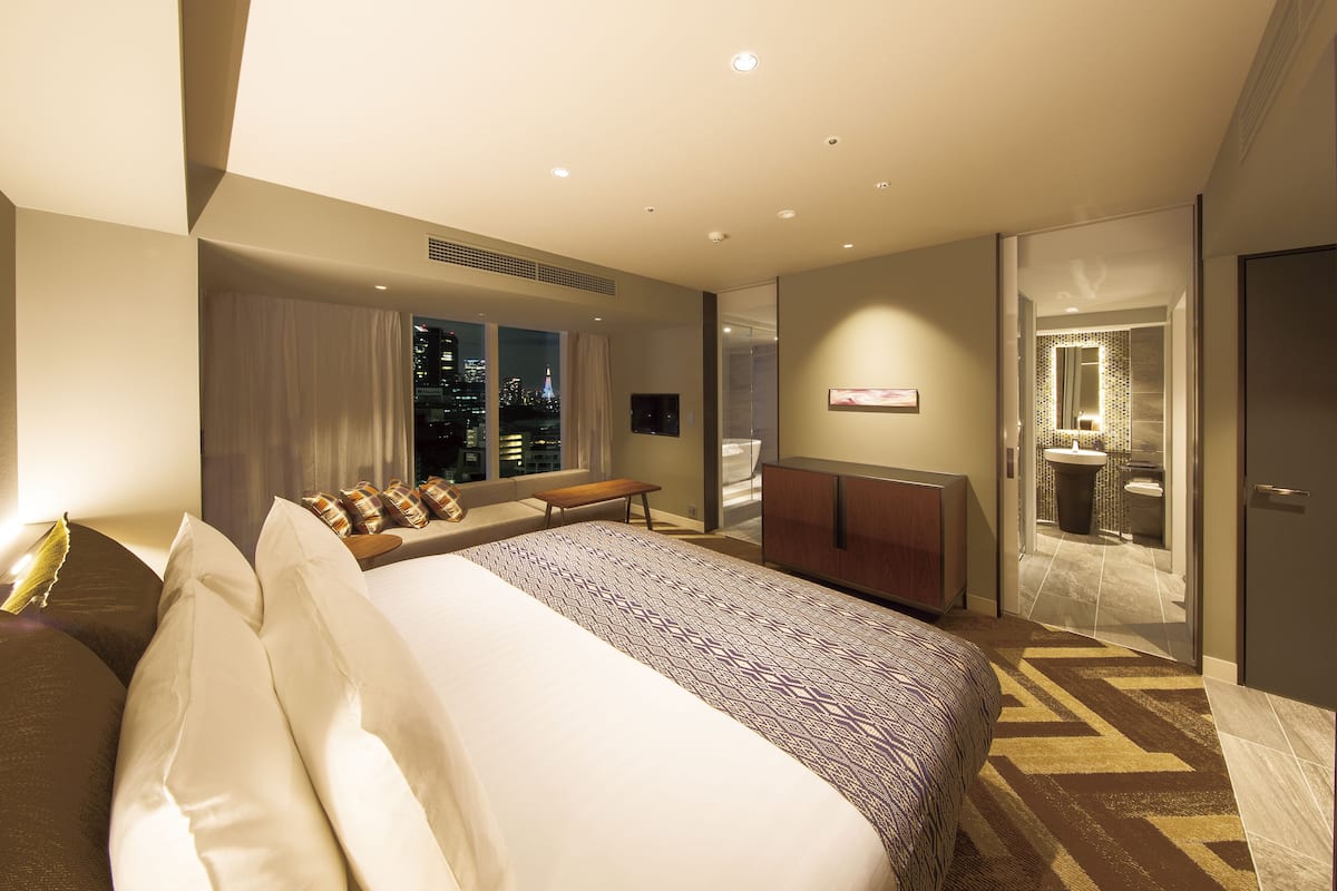 A modern hotel room with a large bed, a desk, and a view of the city at night.