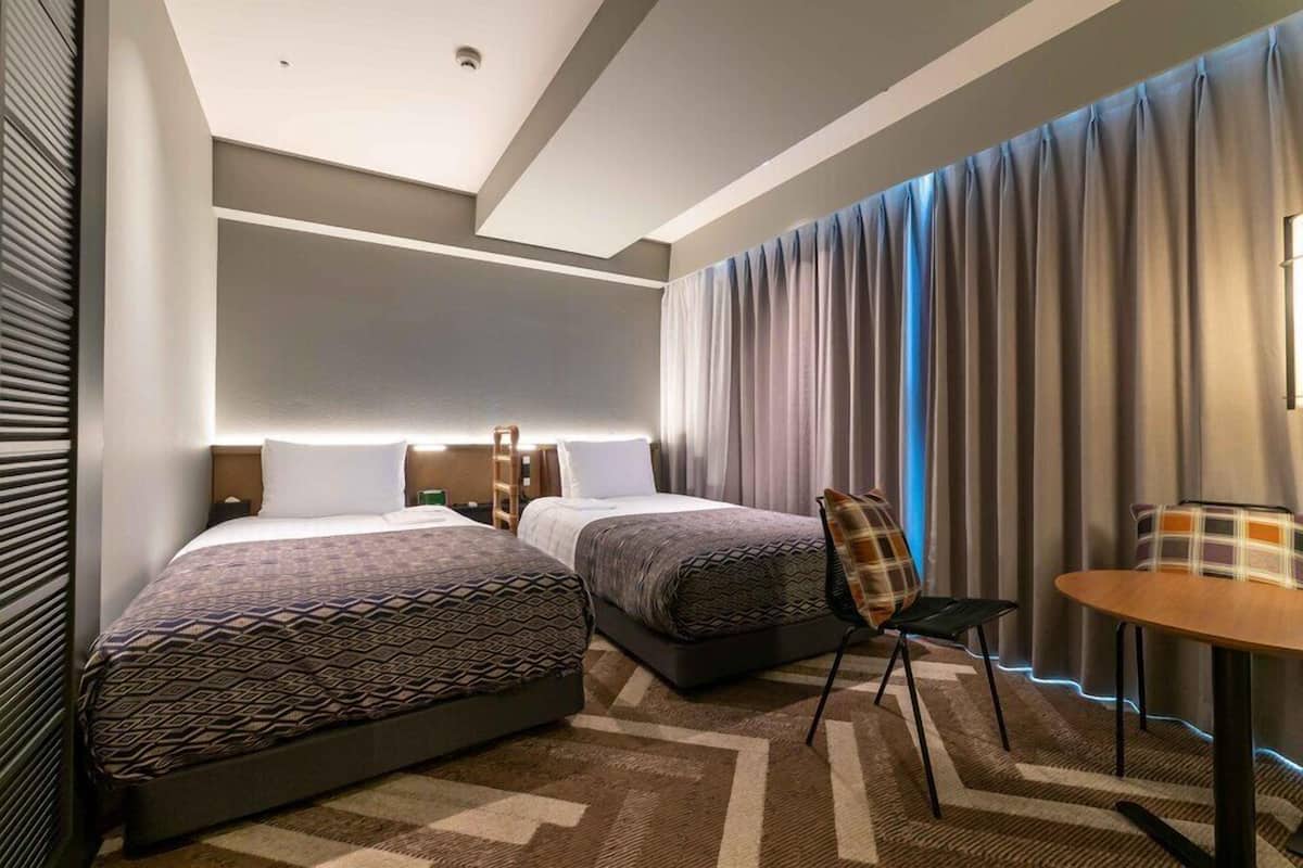 A modern hotel room with two beds, a small table, and a chair.