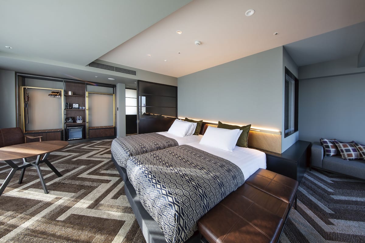 A modern hotel room with two beds, a sofa, a dining table, and a built-in TV.