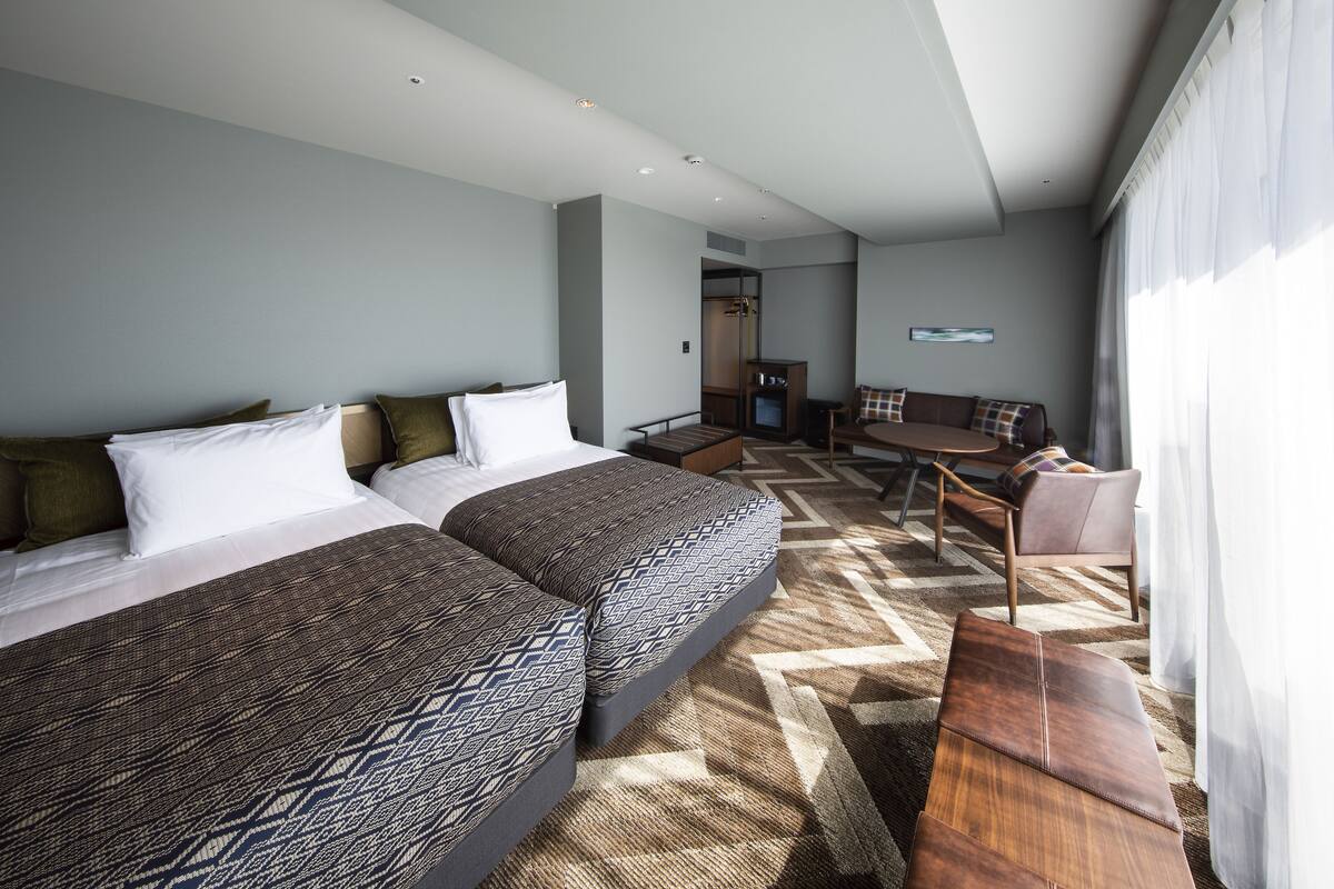 A modern hotel room with two beds, a sofa, a dining area, and a view of the city.