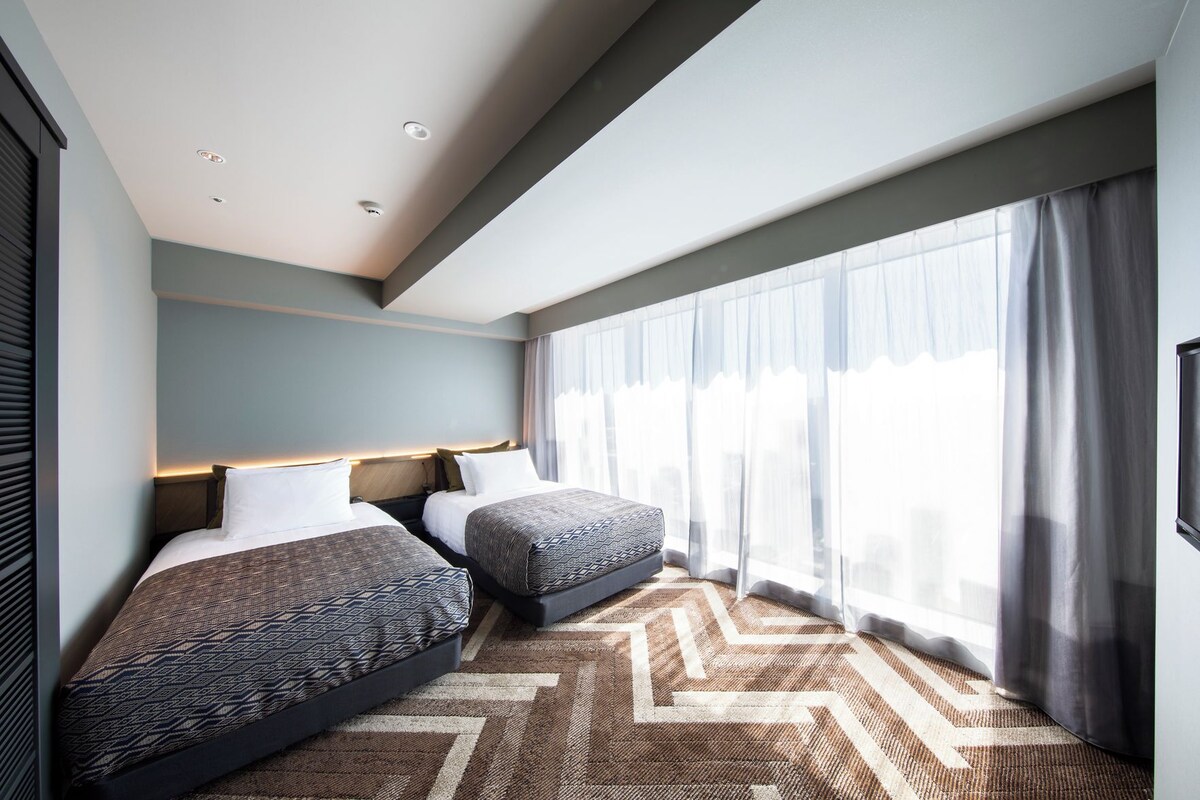 A hotel room with two beds, a large window with sheer curtains, a patterned carpet, and a modern ceiling design.
