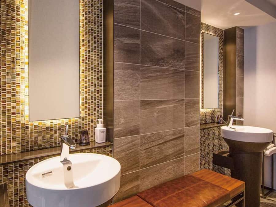 Um banheiro moderno com duas pias, um banco e paredes com mosaicos.