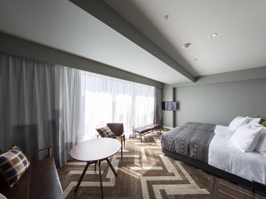 Una habitación de hotel moderna con una cama grande, una mesita pequeña, un sofá y un televisor en la pared.