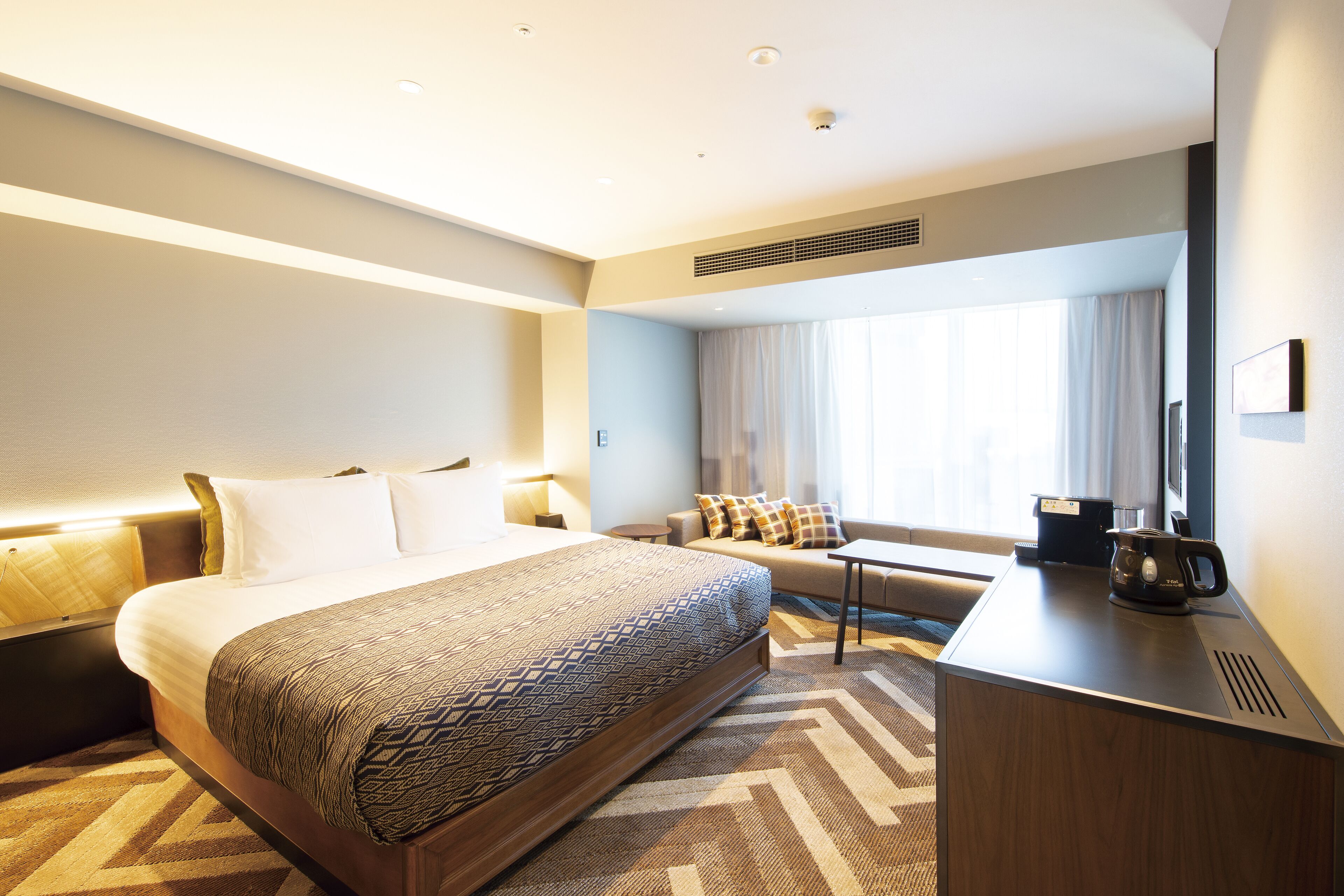 A hotel room with a large bed, a sofa, a desk with a kettle, and a window with curtains.