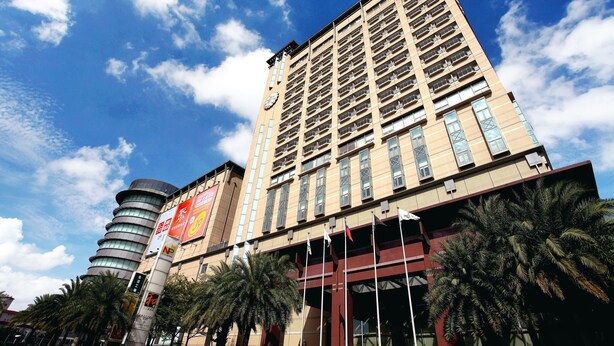 Un hotel de varios pisos con una imponente torre de reloj, fachada moderna y una palmera en primer plano.